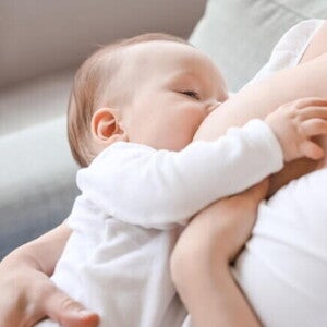 Bebê de roupa branca sendo amamentado pela mãe sentada em um sofá Bebê de roupa branca sendo amamentado pela mãe sentada em um sofá