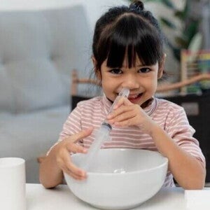 Um bebê deitado com a boca aberta, enquanto uma mão segura uma seringa próxima ao nariz do bebê
