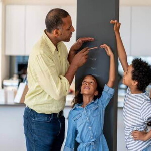 Em uma cozinha, está um homem, uma menina e um menino. A menina está encostada em uma parede, enquanto o homem e o menino marcam na parede qual é a altura dela.