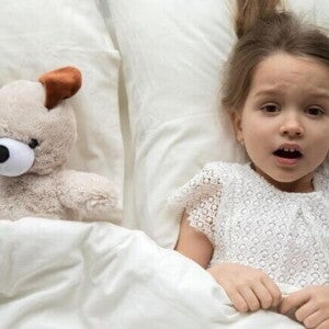 Uma menina assustada está deitada na cama, coberta por um lençol, com expressão de medo, ao lado de um cachorro de pelúcia.