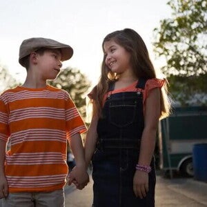 Imagem de um menino e uma menina de mãos dadas se olhando. O menino usa uma boina e uma camisa listrada laranja e branco e a menina está de cabelo solto, usando um macacão escuro e sorrindo Imagem de um menino e uma menina de mãos dadas se olhando. O menino usa uma boina e uma camisa listrada laranja e branco e a menina está de cabelo solto, usando um macacão escuro e sorrindo