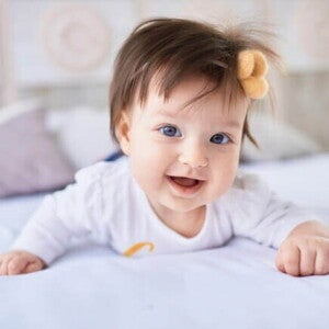 Bebê sorridente deitada de bruços na cama, usando roupa branca e um laço no cabelo