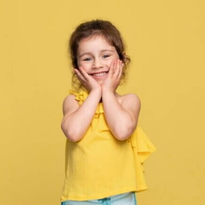 Uma menina sorridente de aproximadamente 3 anos, usando uma blusa amarela, de costas para um fundo amarelo.