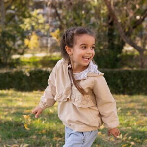 Uma menina de aproximadamente 4 anos brincando ao ar livre em um parque, com um casaco bege e tranças.