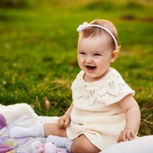 Bebê sorridente usando um vestido claro e uma tiara com flores, sentada em um cobertor sobre a grama.