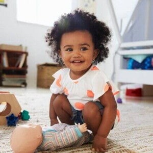 Bebê sorridente de cabelos crespos agachada sobre um tapete, brincando com uma boneca em um ambiente infantil.