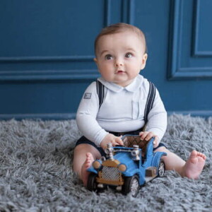 bebê menino sentado em tapete felpudo cinza, com carrinho de brinquedo azul em mãos.