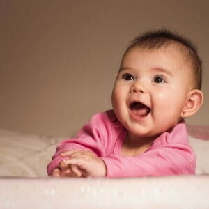 Bebê sorridente deitada de bruços na cama usando uma roupa cor de rosa.
