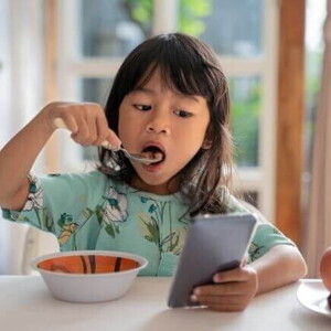 Uma menina come enquanto olha para um celular na mesa.