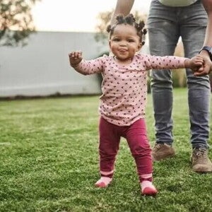 O que é coordenação motora e quais suas fases nas crianças Menina aprendendo a andar e ganhando coordenação motora. Ela caminha no quintal orientada pela mãe, que segura a sua mão esquerda.