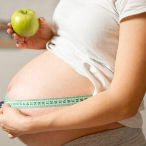 Mulher grávida segurando uma maçã em uma mão e uma fita métrica para costura na outra, ao redor da barriga. 