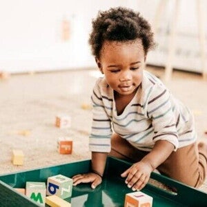 Criança pequena brincando no chão com blocos de montar coloridos em uma bandeja verde, com vários brinquedos espalhados ao redor sobre um tapete bege em um ambiente iluminado. Criança pequena brincando no chão com blocos de montar coloridos em uma bandeja verde, com vários brinquedos espalhados ao redor sobre um tapete bege em um ambiente iluminado.