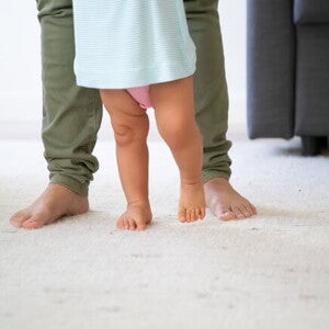 pai, usando calça verde, ajudando o filho a dar os primeiros passos 