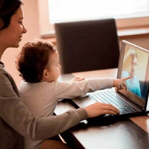 Ilustrando a separação dos pais, uma mãe segura um bebê em seu colo. Eles estão sentados à mesa em frente ao computador. O bebê aponta para a tela, onde há um homem por videochamada. Ilustrando a separação dos pais, uma mãe segura um bebê em seu colo. Eles estão sentados à mesa em frente ao computador. O bebê aponta para a tela, onde há um homem por videochamada.