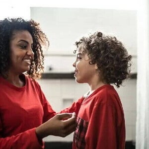 Palavras mágicas Mulher sorrindo com seu filho, ambos usando um pijama vermelho e conversando. Ao fundo, um quarto.