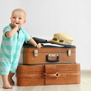 Bebê sorrindo, com a mão na boca, apoiado em malas de viagem.