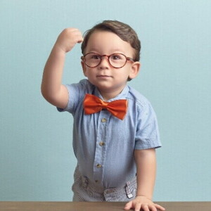 Menino com óculos, gravata vermelha e camisa azul, está apoiado sobre uma mesa, com a mão direita levantada em punho, sob um fundo azul.
