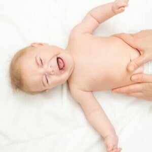Bebê deitado de costas em uma superfície branca, chorando, enquanto um adulto realiza uma massagem suave em sua barriga.