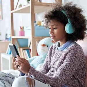 Pré-adolescência #ParaTodosVerem Menina negra pré-adolescente, com cabelo cacheado, solto, usando fones de ouvido azuis enquanto olha a tela do celular. Ela está sentada em uma cama com almofadas e veste blusão com calça jeans. O fundo da foto é um quarto de menina.