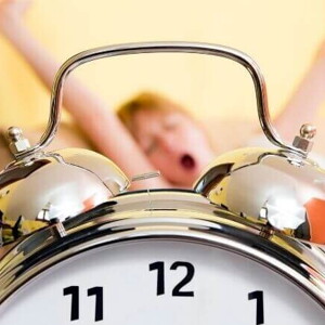 Relógio despertador prateado em primeiro plano, com uma criança ao fundo bocejando e se espreguiçando em uma cama Relógio despertador prateado em primeiro plano, com uma criança ao fundo bocejando e se espreguiçando em uma cama