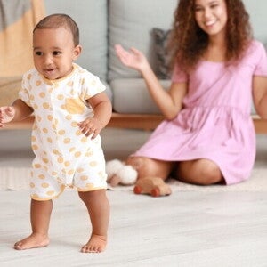 Um bebê sorrindo e em pé no chão, olhando para a frente, com uma mulher sentada no chão ao fundo, sorrindo. Um bebê sorrindo e em pé no chão, olhando para a frente, com uma mulher sentada no chão ao fundo, sorrindo.