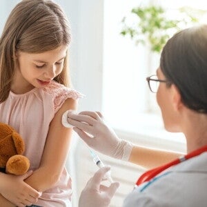 Uma menina segura um ursinho de pelúcia enquanto uma profissional de saúde, prepara o braço da menina para uma injeção com um algodão. Uma menina segura um ursinho de pelúcia enquanto uma profissional de saúde, prepara o braço da menina para uma injeção com um algodão.
