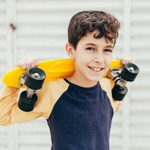 Menino sorrindo e segurando um skate pequeno em cima dos ombros