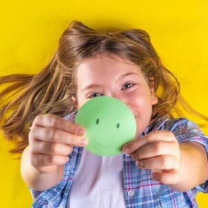 Uma criança deitada e sorrindo, segurando um círculo de papel verde com um rosto sorridente desenhado.