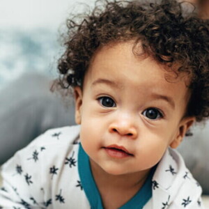 Bebê com cabelo cacheado vestindo um pijama branco com detalhes azuis, olhando diretamente para a câmera.