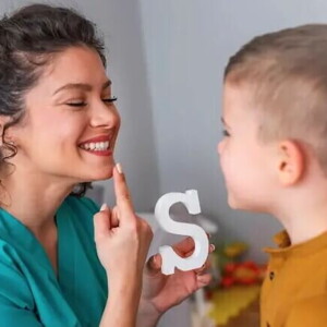 Uma mulher sorrindo, apontando para a própria boca e segurando um objeto em forma de letra “S”, enquanto uma criança observa e tenta imitar a ação da mulher.