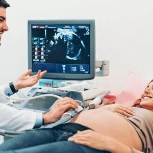 Mulher grávida deitada em uma maca durante exame de ultrassom, enquanto médico realiza o procedimento e conversa com a paciente; ao fundo, monitor exibe a imagem do bebê. Mulher grávida deitada em uma maca durante exame de ultrassom, enquanto médico realiza o procedimento e conversa com a paciente; ao fundo, monitor exibe a imagem do bebê.