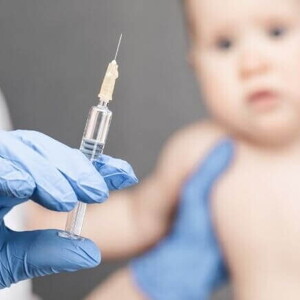 Mão com luva cirúrgica segurando seringa com a vacina da coqueluche, enquanto bebê aguarda a aplicação Mão com luva cirúrgica segurando seringa com a vacina da coqueluche, enquanto bebê aguarda a aplicação