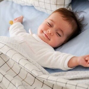 Bebê dormindo de barriga para cima em uma cama com lençóis e cobertor, transmitindo sensação de conforto e tranquilidade.