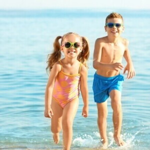 Duas crianças sorrindo e correndo na praia, usando óculos de sol e roupa de banho.