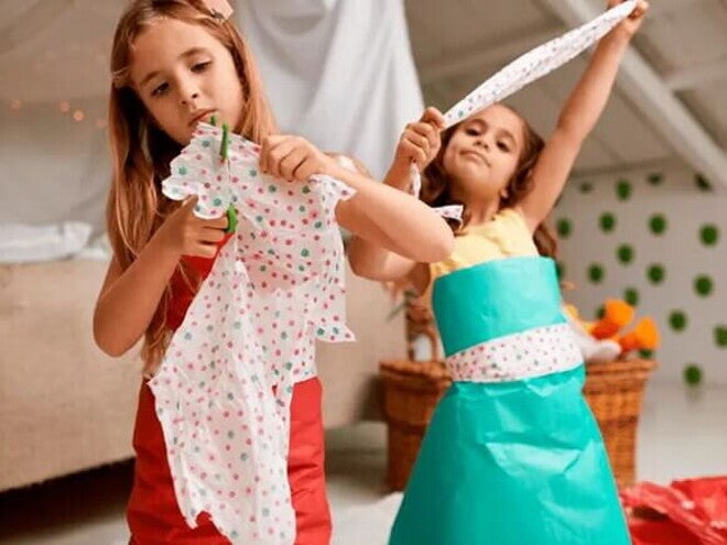 Duas crianças de cerca de 8 anos cortam papel colorido com uma tesoura sem ponta customizando pequenas peças de roupa com este material. Uma delas está utilizando, sobre a sua roupa comum, um vestido feito de papel. Duas crianças de cerca de 8 anos cortam papel colorido com uma tesoura sem ponta customizando pequenas peças de roupa com este material. Uma delas está utilizando, sobre a sua roupa comum, um vestido feito de papel.