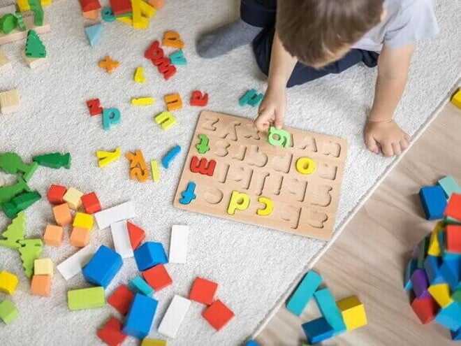Em um piso de madeira com um tapete cinza, está sentado um menino,vestindo uma camiseta branca. Ele está brincando com um tabuleiro de encaixar letras, enquanto segurando uma delas. Há letras e outras peças coloridas espalhadas ao seu redor. Em um piso de madeira com um tapete cinza, está sentado um menino,vestindo uma camiseta branca. Ele está brincando com um tabuleiro de encaixar letras, enquanto segurando uma delas. Há letras e outras peças coloridas espalhadas ao seu redor.