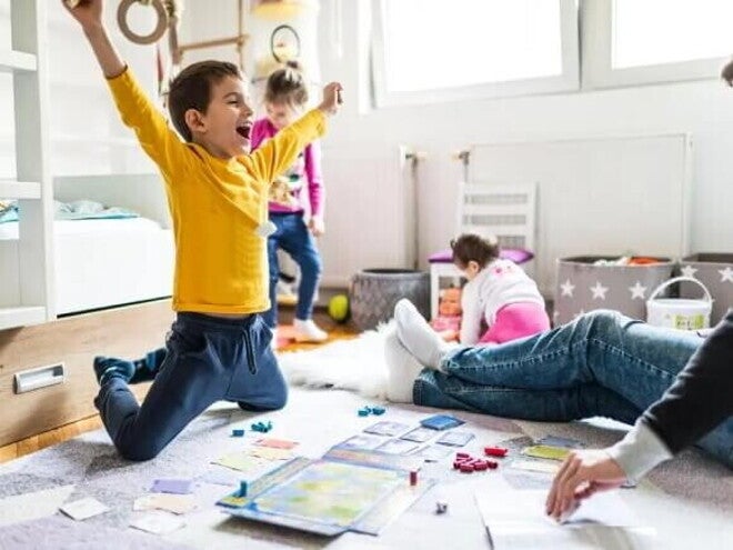 16 atividades lúdicas para fazer em casa com as crianças Sala com um adulto, onde só é possível ver suas pernas usando calça jeans e meia. Na mesma sala, estão três crianças. Uma garota com cerca de 1 ano de idade em segundo plano, branca, de cabelos curtos, de costas brincando com uma boneca. Outra garota com cerca de quatro anos, branca, cabelos loiros presos, calça jeans e blusa de manga comprida. Ela parece estar feliz dançando. A criança em destaque é um menino, branco, com cabelos curtos, de braços abertos comemorando uma vitória em um jogo