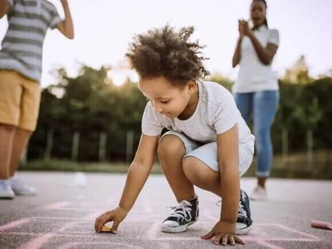 Imagem de uma criança ao centro brincando de uma brincadeira africana no chão. Ao fundo seus pais torcem por ela Imagem de uma criança ao centro brincando de uma brincadeira africana no chão. Ao fundo seus pais torcem por ela
