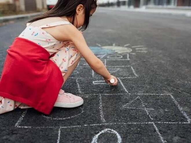 Uma menina de vestido e tênis está agachada na rua desenhando uma amarelinha no chão. A foto faz alusão ao tema de brincadeiras de rua Uma menina de vestido e tênis está agachada na rua desenhando uma amarelinha no chão. A foto faz alusão ao tema de brincadeiras de rua