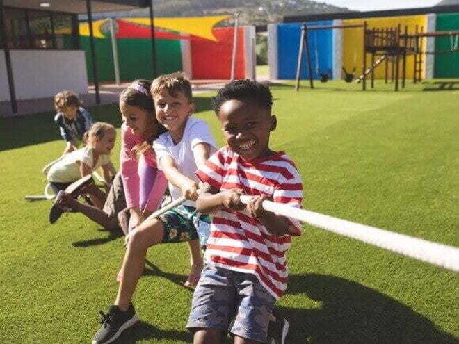 Como passar as brincadeiras do folclore brasileiro para seus filhos? Em um parque ao ar livre, três meninos e duas meninas estão se divertindo em uma brincadeira de cabo de guerra. As crianças, sorridentes, estão puxando a corda.