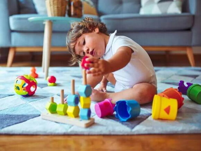 Um bebê está sentado no tapete de uma sala de estar mexendo com brinquedos de empilhar e montar, que é uma das brincadeiras que estimulam o desenvolvimento infantil Um bebê está sentado no tapete de uma sala de estar mexendo com brinquedos de empilhar e montar, que é uma das brincadeiras que estimulam o desenvolvimento infantil