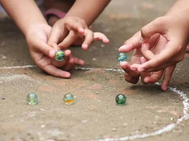 Duas crianças brincam de bola de gude no chão em um círculo desenhado de giz. A foto faz alusão ao tema de brincadeiras indígenas Duas crianças brincam de bola de gude no chão em um círculo desenhado de giz. A foto faz alusão ao tema de brincadeiras indígenas