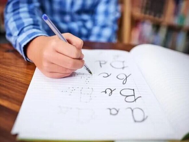 Imagem de um menino segurando uma caneta azul. Em um caderno ele vai ligando os pontinhos para formar as palavras: a,b,c e d. A foto faz alusão ao tema caligrafia. Imagem de um menino segurando uma caneta azul. Em um caderno ele vai ligando os pontinhos para formar as palavras: a,b,c e d. A foto faz alusão ao tema caligrafia.