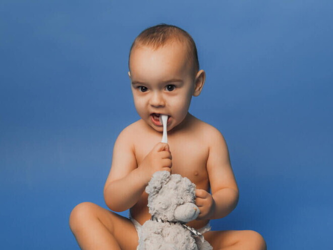 Bebê sentado com uma escova de dentes na boca, segurando um urso de pelúcia Bebê sentado com uma escova de dentes na boca, segurando um urso de pelúcia