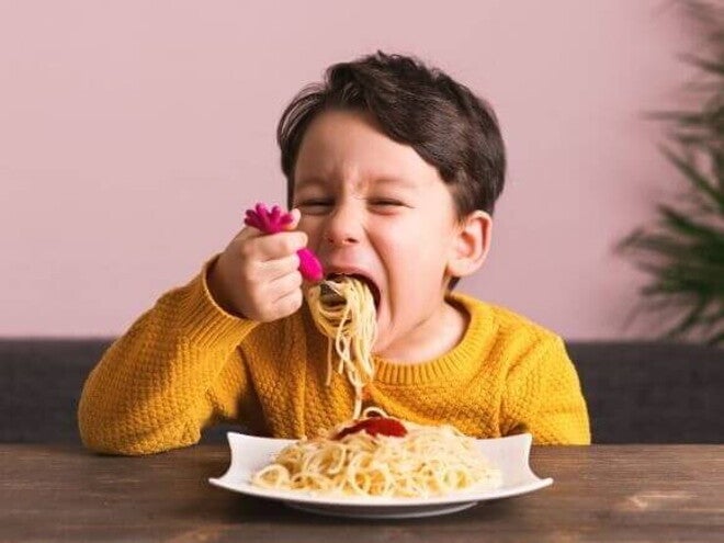 Criança sentada à mesa, comendo um prato de macarrão, em ambiente interno com parede rosa e planta ao fundo. Criança sentada à mesa, comendo um prato de macarrão, em ambiente interno com parede rosa e planta ao fundo.
