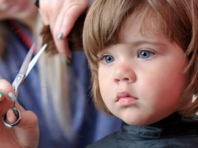 Uma menina recebendo um corte de cabelo. Ao seu lado, a mão de uma mulher segura uma mecha do seu cabelo, enquanto outra mão segura uma tesoura pequena Uma menina recebendo um corte de cabelo. Ao seu lado, a mão de uma mulher segura uma mecha do seu cabelo, enquanto outra mão segura uma tesoura pequena