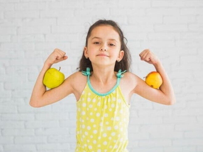 Menina sorridente fazendo pose de força com os braços flexionados, segurando uma maçã em cada braço, em frente a uma parede branca. Menina sorridente fazendo pose de força com os braços flexionados, segurando uma maçã em cada braço, em frente a uma parede branca.