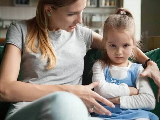 Mãe e filha sentadas no sofá da sala, mãe abraça a criança e conversa, criança parece estressada com os braços cruzados e olhar distante Mãe e filha sentadas no sofá da sala, mãe abraça a criança e conversa, criança parece estressada com os braços cruzados e olhar distante