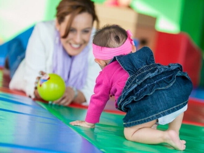 Um bebê engatinhando em direção a uma mulher sorridente, que observa o bebê com uma bola verde na mão. Um bebê engatinhando em direção a uma mulher sorridente, que observa o bebê com uma bola verde na mão.
