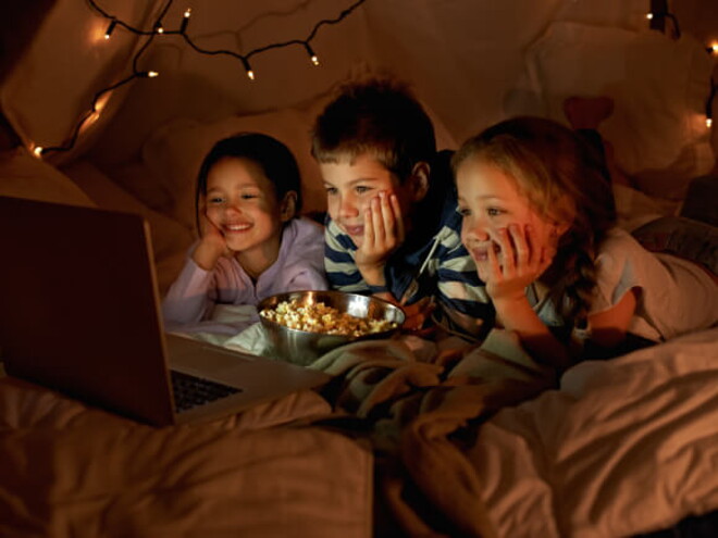 Três crianças, sendo um menino e duas meninas, deitados assistindo no notebook enquanto comem pipoca. Ao fundo, luminárias em uma cabana. Três crianças, sendo um menino e duas meninas, deitados assistindo no notebook enquanto comem pipoca. Ao fundo, luminárias em uma cabana.